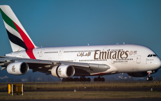 Un Airbus A380 d'Emirates, une création de la célèbre compagnie aérienne, roule sur une piste. L'avion brille de blanc, orné du logo Emirates et d'une livrée mettant en valeur les couleurs du drapeau des Émirats arabes unis sur sa queue. Un ciel bleu clair et un paysage lointain offrent une toile de fond époustouflante.