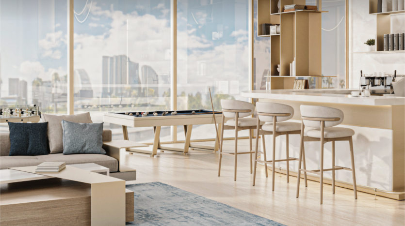 Un espace de vie moderne et lumineux de la collection Cove Edition 2 avec de grandes fenêtres présentant un paysage urbain. La pièce comprend un baby-foot, un coin bar avec des tabourets, un canapé gris confortable et une table basse en bois sur un tapis bleu clair. La décoration est minimaliste avec une palette de couleurs neutres.