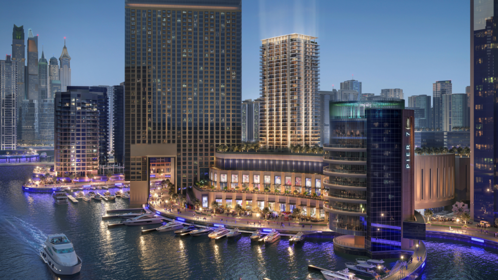 La nuit, le paysage urbain est animé et les immeubles hauts et illuminés bordent le front de mer de Marina Cove. Les bateaux sont amarrés à la marina et la zone regorge d'activité. L'horizon est visible en arrière-plan sous un ciel clair et sombre.