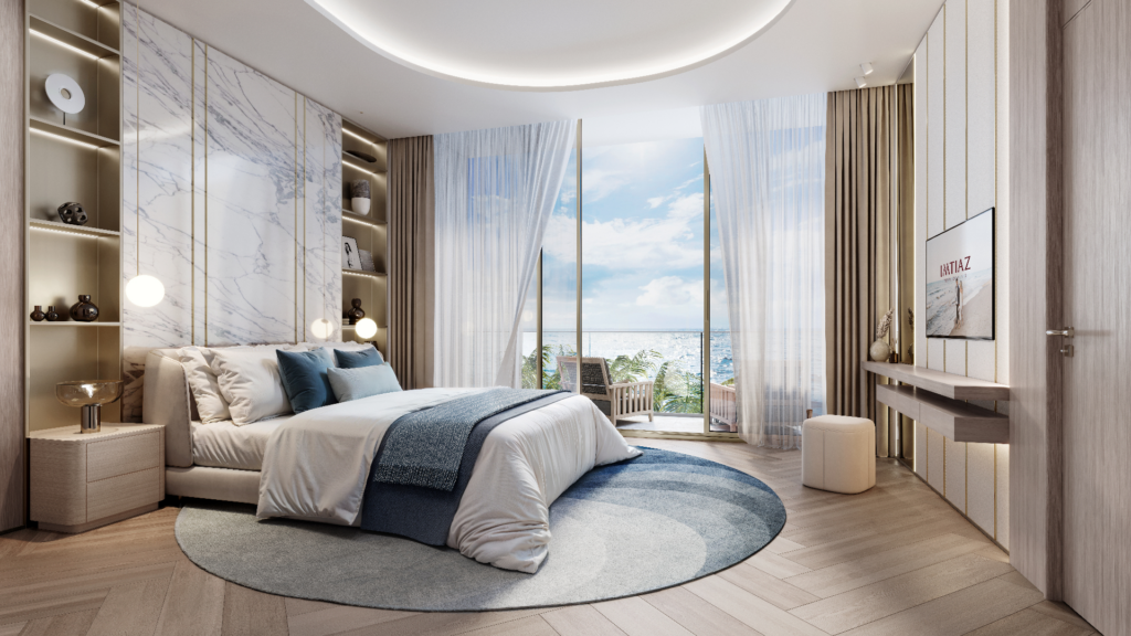 Une chambre moderne avec un grand lit, un tapis rond et un mur d'accent en marbre. Les baies vitrées révèlent une vue sur la mer et un balcon. Les tons neutres et le mobilier élégant créent une atmosphère sereine. Une télévision murale est visible sur la droite.