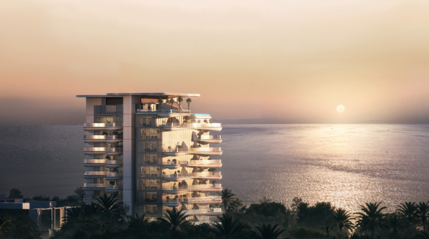 Un bâtiment moderne à plusieurs étages avec balcons, connu sous le nom de Beach Walk Grand, surplombe un océan serein au coucher du soleil. Le ciel est d'un orange chaleureux et le soleil projette une lueur réfléchissante sur l'eau calme, tandis que les palmiers se balancent doucement au premier plan.