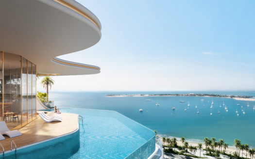 Modernes Dach mit Infinity-Pool, der einen Panoramablick auf den Ozean im Beach Walk Grand bietet. Eine Person räkelt sich auf einem Stuhl neben den Panoramafenstern. Segelboote säumen das blaue Meer, darunter ein Sandstrand und Palmen unter einem hellblauen Himmel.
