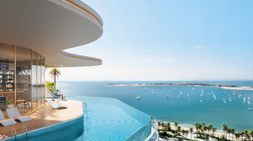 Toit moderne avec piscine à débordement donnant sur une vue panoramique sur l'océan au Beach Walk Grand. Une personne se prélasse sur une chaise près des baies vitrées. Des voiliers parsèment la mer bleue, avec une plage de sable et des palmiers en contrebas sous un ciel bleu clair.