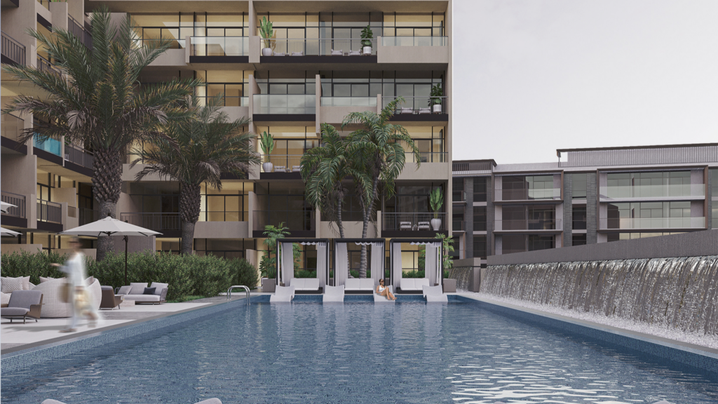 A modern apartment complex with a large swimming pool in the foreground, lined with sun loungers and palm trees. A waterfall runs along the right-hand side. Apartment balconies overlook the pool.