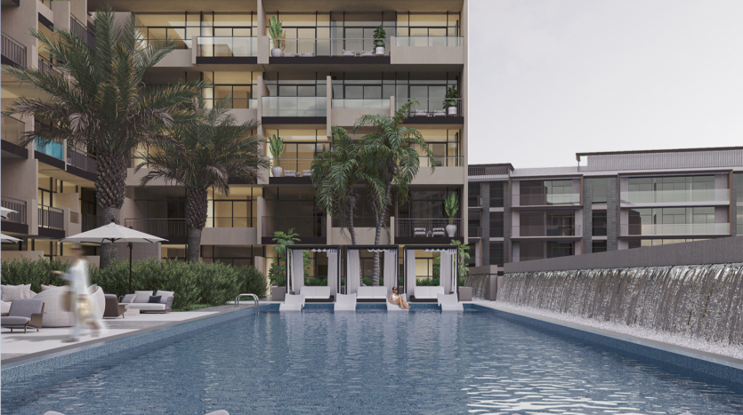 A modern apartment complex with a large swimming pool in the foreground, lined with sun loungers and palm trees. A waterfall runs along the right-hand side. Apartment balconies overlook the pool.