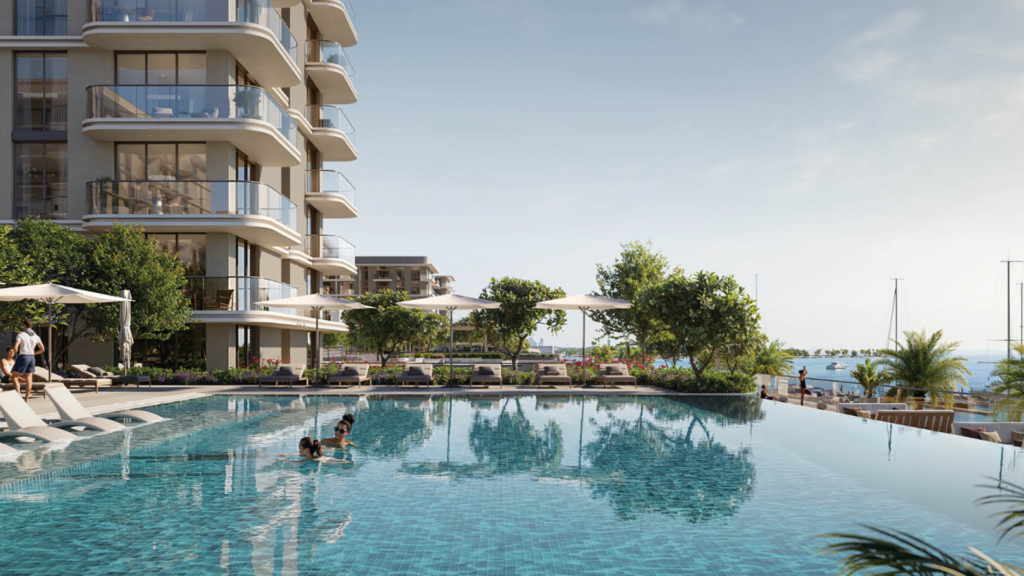 Marina Place 1 propose un immeuble d'appartements moderne avec des balcons en verre donnant sur une piscine à débordement dans laquelle une personne nage. Des chaises longues et des parasols au bord de la piscine bordent le patio, avec une végétation luxuriante et une marina en arrière-plan sous un ciel clair.