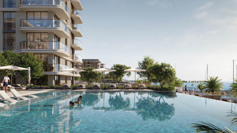 Marina Place 1 propose un immeuble d'appartements moderne avec des balcons en verre donnant sur une piscine à débordement dans laquelle une personne nage. Des chaises longues et des parasols au bord de la piscine bordent le patio, avec une végétation luxuriante et une marina en arrière-plan sous un ciel clair.