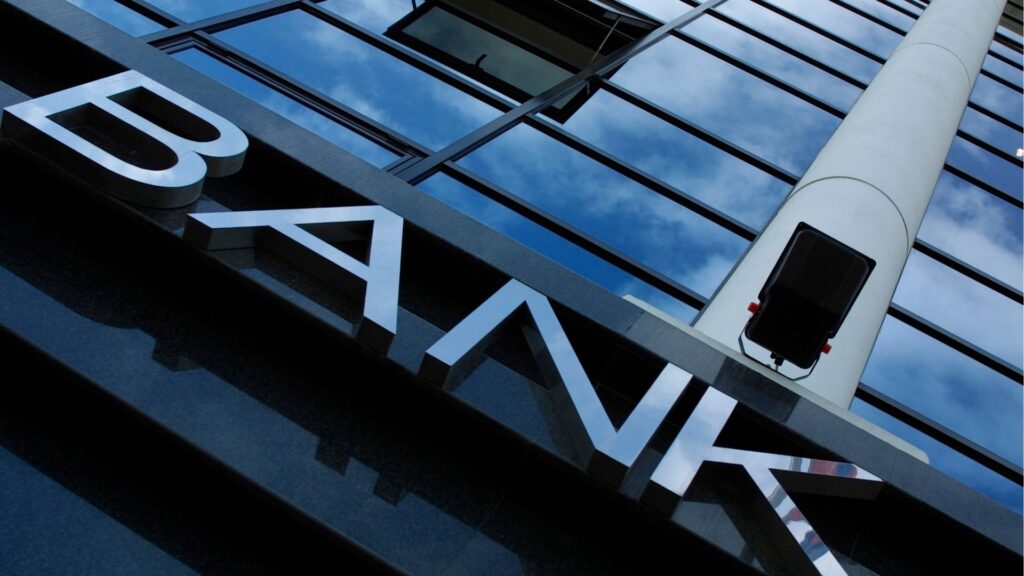 Nahaufnahme eines modernen Gebäudes mit großen verspiegelten Fenstern, die einen Teil eines Schildes zeigen, auf dem in Metallbuchstaben "BANK" steht - ideal für diejenigen, die sich fragen, wie man in Dubai einen Hauskredit aufnimmt. Der Himmel und die Wolken spiegeln sich in den Fenstern.