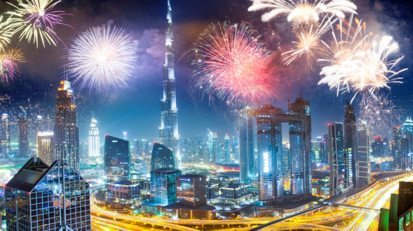 Un paysage urbain animé la nuit avec un feu d'artifice coloré illuminant le ciel de Dubaï. Les gratte-ciels sont illuminés et les routes très fréquentées sont visibles en contrebas, avec des traînées lumineuses provenant des véhicules en mouvement. C'est un guide parfait pour les activités hivernales dans cette ville dynamique.