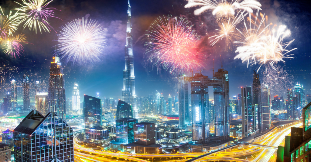 Nachts wird die Skyline von Dubai von einem farbenfrohen Feuerwerk erhellt. Hohe Wolkenkratzer, darunter ein prominenter Turm, sind zwischen den Lichtern zu sehen, während sich die Straßen durch die Szenerie schlängeln und ein Gefühl von Feierlichkeit und Energie erzeugen, das einem Neujahrsfest ähnelt.