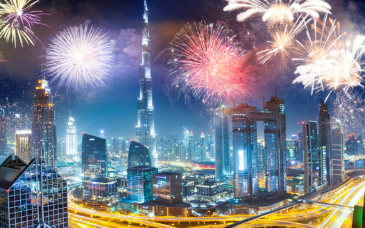 La nuit, l'horizon de Dubaï est illuminé par un feu d'artifice coloré. De hauts gratte-ciels, dont une tour proéminente, sont visibles parmi les lumières, tandis que les routes serpentent à travers la scène, créant un sentiment de fête et d'énergie proche d'un festival de nouvel an.