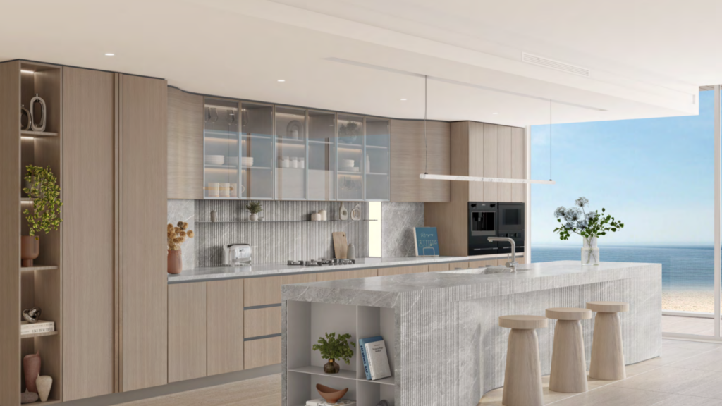 Cuisine moderne à Ellington Cove avec des meubles en bois clair et un grand îlot en pierre. Les armoires supérieures vitrées présentent la vaisselle, tandis que les appareils intégrés et les étagères ouvertes abritent les plantes et la décoration. Vue sur l'océan à travers une fenêtre spacieuse avec trois tabourets en bois près de l'îlot.