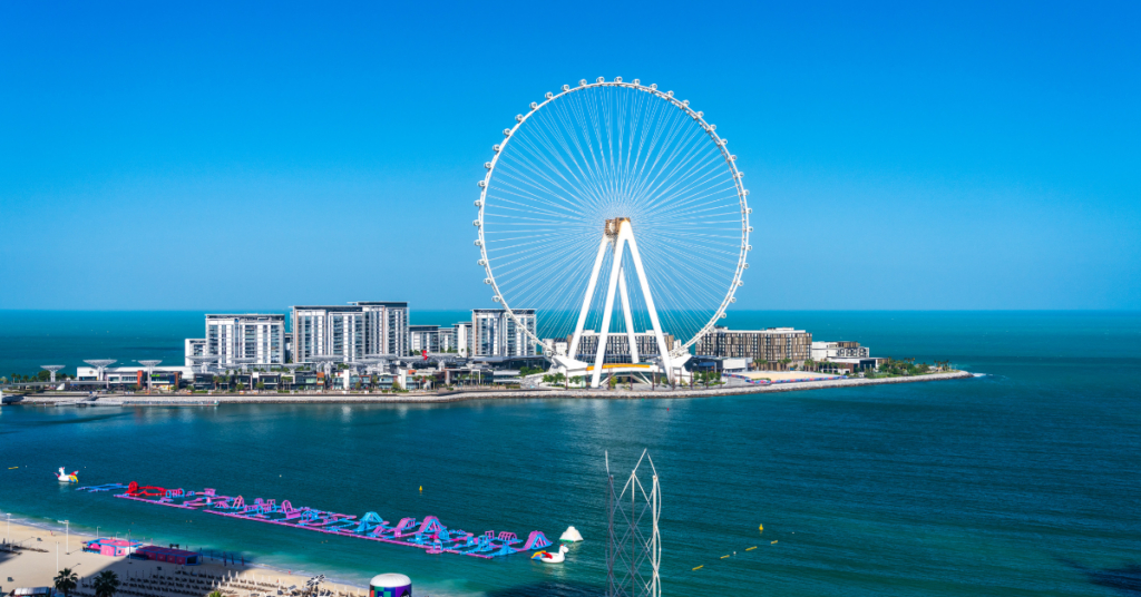 Une scène côtière avec une grande roue sur une île, entourée de plusieurs bâtiments blancs, offre une évasion pittoresque des activités hivernales à Dubaï. La mer calme et bleue s'étend au premier plan avec un parc aquatique flottant près du rivage sous un ciel bleu clair.