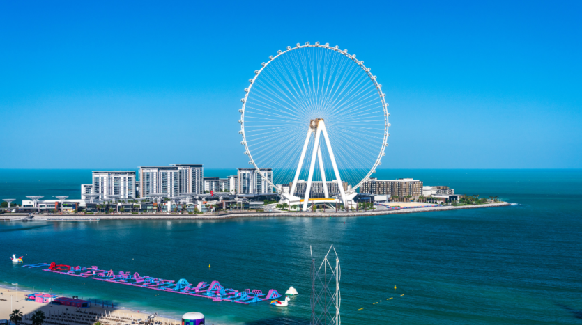 Une scène côtière avec une grande roue sur une île, entourée de plusieurs bâtiments blancs, offre une évasion pittoresque des activités hivernales à Dubaï. La mer calme et bleue s'étend au premier plan avec un parc aquatique flottant près du rivage sous un ciel bleu clair.