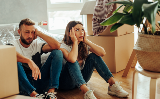 Un homme et une femme sont assis par terre, l'air fatigué et stressé, entourés de cartons de déménagement dans une pièce ensoleillée. Alors qu'ils réfléchissent à la façon de meubler un appartement à Dubaï, une grande plante en pot se détache au premier plan, offrant un soupçon de sérénité au milieu du chaos.