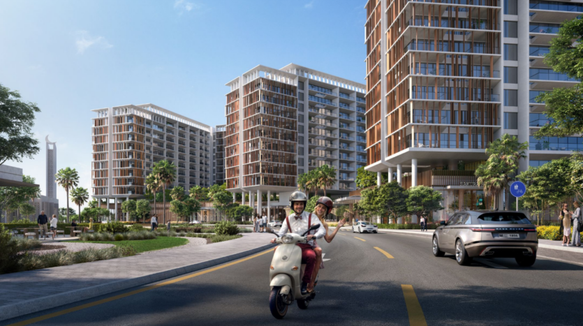 Un couple conduit un scooter Parkfive dans une rue de la ville avec des immeubles modernes en arrière-plan. Une voiture suit derrière, et il y a des arbres et de la verdure le long de la route. Le ciel est clair et ensoleillé.