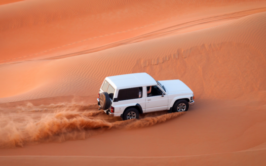 Un SUV blanc, tel un guide silencieux, traverse de vastes dunes de sable rouge, projetant derrière lui un panache de sable sous le ciel dégagé. La scène capture le mouvement dynamique et la vaste étendue du paysage désertique de Dubaï.