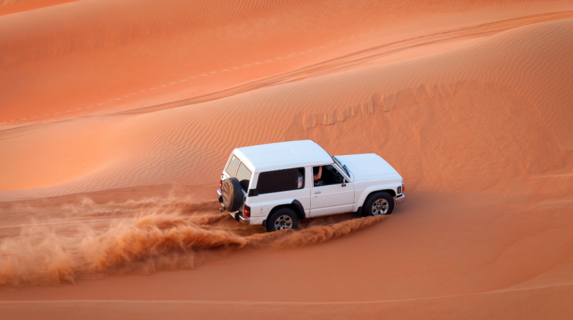 Un SUV blanc, tel un guide silencieux, traverse de vastes dunes de sable rouge, projetant derrière lui un panache de sable sous le ciel dégagé. La scène capture le mouvement dynamique et la vaste étendue du paysage désertique de Dubaï.