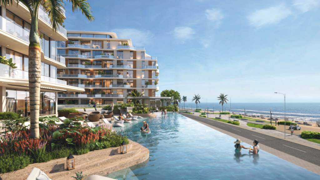Modern beachfront resort with a luxurious infinity pool surrounded by tropical plants. Guests relax in the pool and nearby seating areas. The building has large balconies and the ocean is visible under a clear blue sky.
