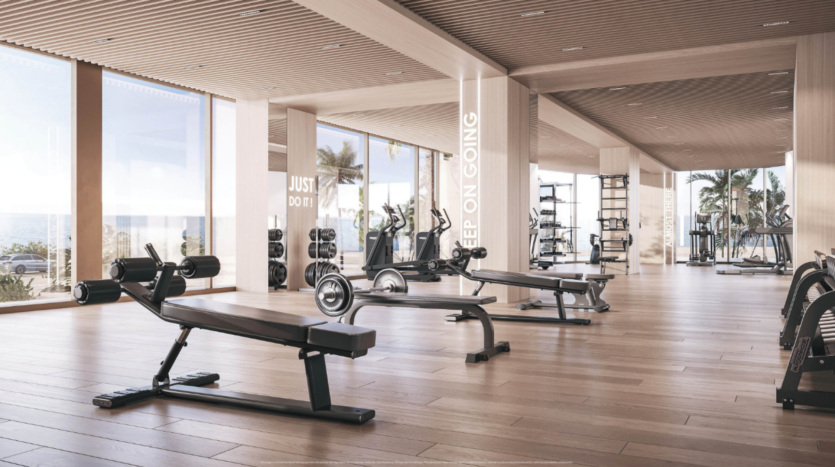 A modern gym with parquet flooring, equipped with weight benches, exercise bikes and a variety of gym equipment. Large windows offer views of palm trees and the ocean. Motivational phrases are displayed on the mirrors.