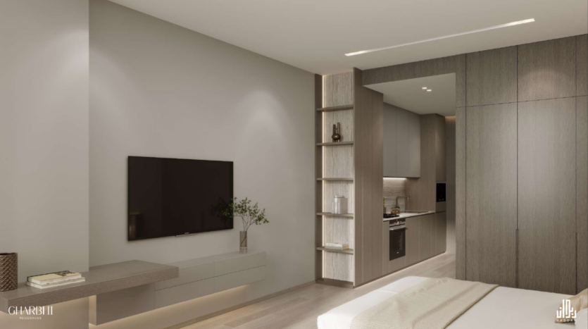 Una moderna camera d'albergo dal design minimalista presenta un televisore a parete di fronte al letto, un'elegante scrivania e scaffali aperti. La parete di fondo rivela un angolo cottura compatto con fornelli e lavello. I toni neutri e l'illuminazione discreta creano un ambiente caldo.