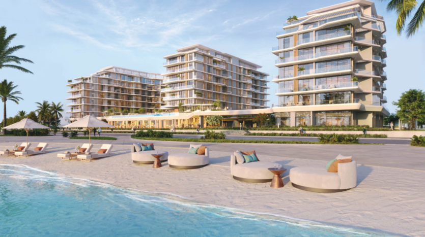 Seafront view of modern luxury apartments with glass facades, surrounded by palm trees. The sandy beach in the foreground features sun loungers and round tables, with a clear blue sky above.
