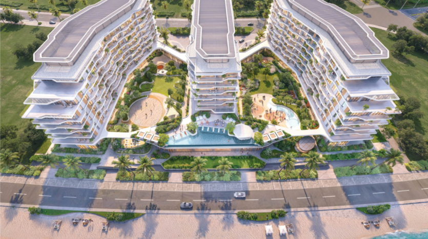 Aerial view of a modern, multi-storey beachfront hotel with two curved buildings. The hotel features terraces, lush gardens, a central swimming pool and palm trees lining the entrance. The beach and a road are visible in the foreground.