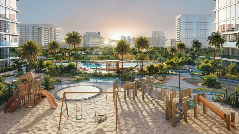 Panoramic view of Verano, a modern residential complex with a playground in the foreground, complete with swings and slides. In the background, palm trees, swimming pools, gardens and tall buildings are illuminated by a setting sun.