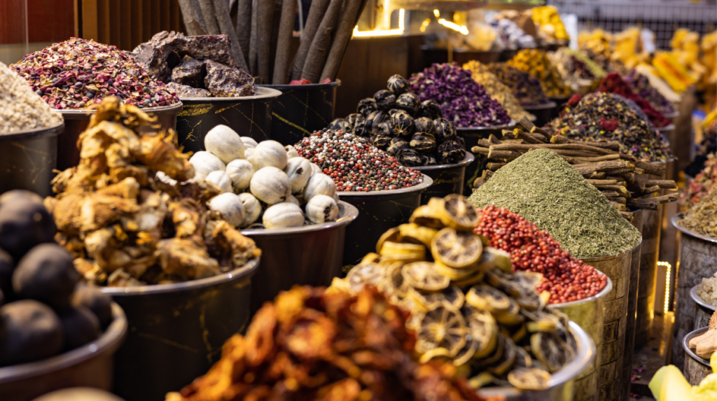 Un'esposizione colorata di varie spezie e prodotti essiccati in grandi contenitori di metallo in una bancarella del mercato di Dubai. L'assortimento comprende erbe, frutta secca, semi e spezie, creando una scena aromatica che riflette le ricche pratiche culturali di Dubai.