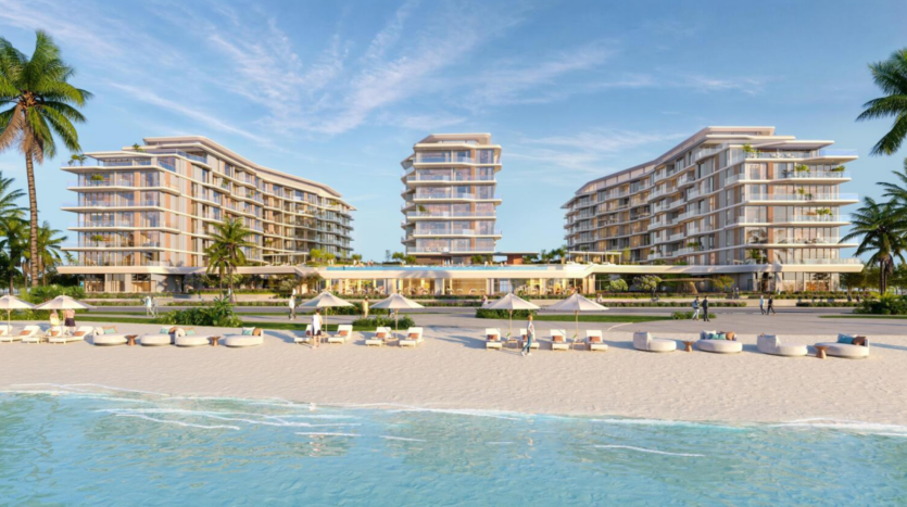 A beach resort consisting of two modern multi-storey buildings surrounded by palm trees. White umbrellas and sun loungers line the sandy shore, with the calm blue waves of the ocean in the foreground under a clear, sunny sky.