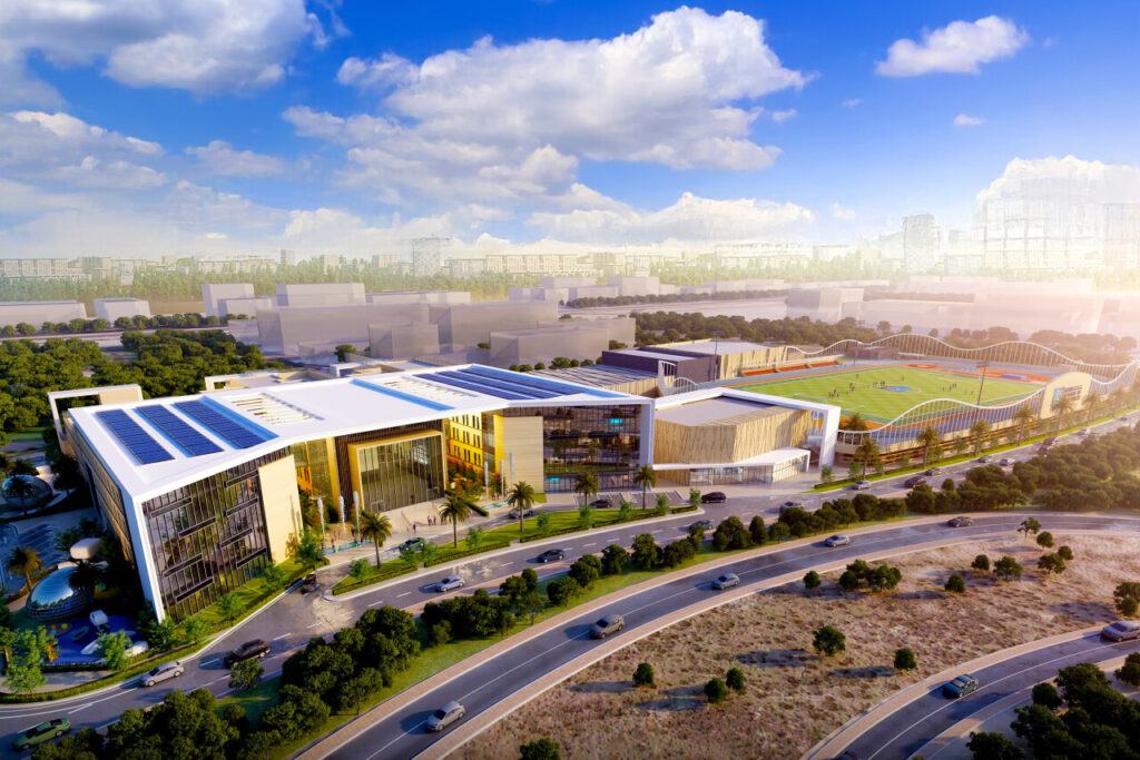 Aus der Luft betrachtet entfaltet sich Dubai Sports City mit modernen Komplexen, die mit Solarzellen geschmückt und von Bäumen umgeben sind. Eine Schule ist in der Nähe eingebettet, während ein Sportplatz mit Tribünen auffällt. Die Straßen beleben sich im Vordergrund vor dem Hintergrund des Horizonts unter einem teilweise bewölkten Himmel.
