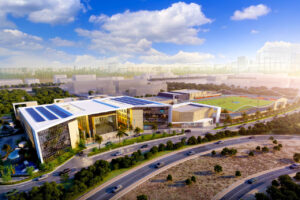 Dall'alto, Dubai Sports City si presenta con moderni complessi ornati da pannelli solari e circondati da alberi. Nelle vicinanze si trova una scuola, mentre spicca un campo sportivo con gradinate. Le strade prendono vita in primo piano contro l'orizzonte, sotto un cielo parzialmente nuvoloso.