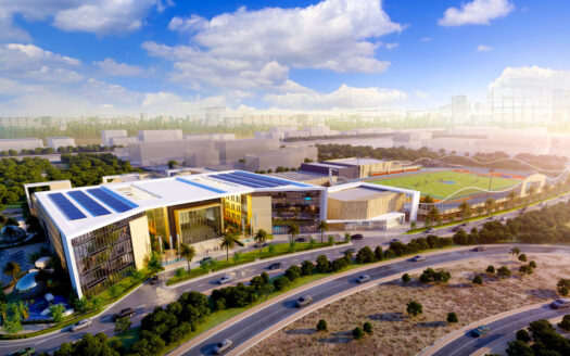 Desde el aire, la Ciudad Deportiva de Dubai se despliega con modernos complejos adornados con paneles solares y rodeados de árboles. En las inmediaciones hay una escuela y destaca un campo de deportes con gradas. Las carreteras cobran vida en primer plano contra el horizonte bajo un cielo parcialmente nublado.