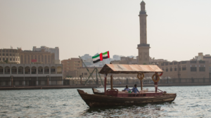 Un tradizionale dhow di legno naviga sul torrente, con la bandiera degli Emirati Arabi Uniti che sventola sopra. Sullo sfondo, il paesaggio urbano di Dubai è costituito da imponenti edifici e da un alto minareto, emblema del cielo limpido.