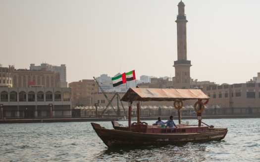 Eine traditionelle hölzerne Dhow segelt auf dem Creek, über ihr weht die Flagge der Vereinigten Arabischen Emirate. Im Hintergrund ist die Stadtlandschaft von Dubai zu sehen, die aus imposanten Gebäuden und einem hohen Minarett besteht, die sinnbildlich für den klaren Himmel stehen.