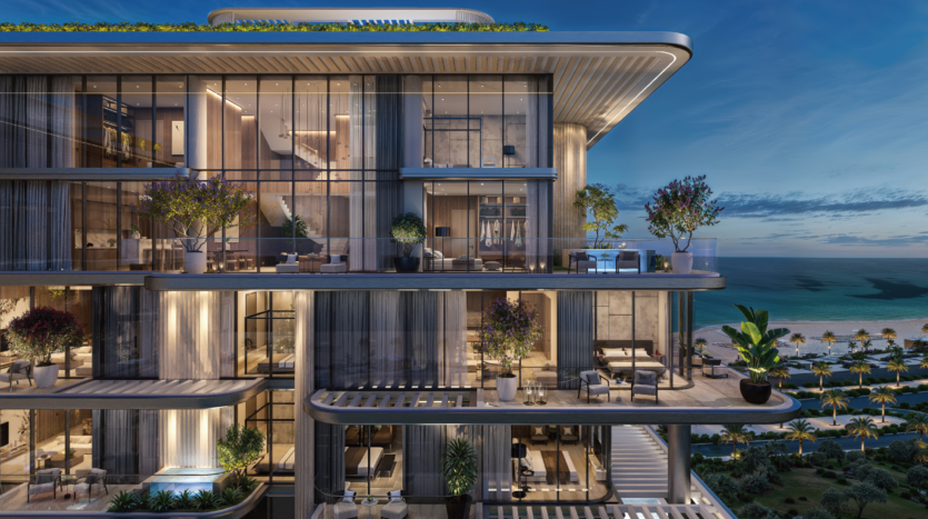 Moderno edificio a più piani con ampi balconi in vetro decorati con piante e arredi. La struttura si affaccia su una spiaggia e sull'oceano, sullo sfondo di un cielo crepuscolare. Gli interni sono illuminati e rivelano elementi di design contemporaneo.
