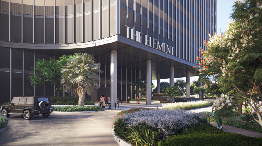 L'elegante ingresso di "The Element" è caratterizzato da grandi vetrate e colonne eleganti. Le persone si riuniscono tra il verde lussureggiante, gli alberi e un SUV nero parcheggiato nel vialetto, che riflette l'estetica moderna di Sobha One.