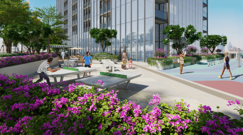 Le persone si godono le attività all'aperto sulla moderna terrazza sul tetto del Sobha One, con tavoli da ping pong, aree per sedersi e una vegetazione lussureggiante. Lo spazio è circondato da fiori viola e offre una vista sui vicini edifici della città sotto un cielo azzurro e limpido.