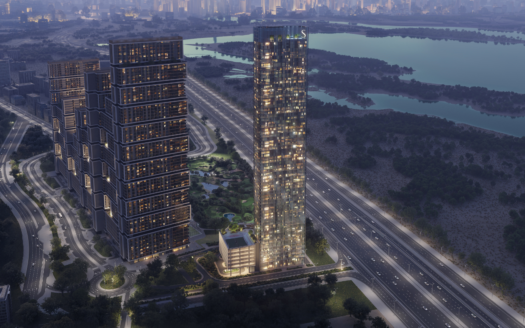 Vue aérienne de deux gratte-ciel modernes illuminés la nuit, probablement le complexe Sobha One, entouré de routes à la circulation fluide. Un paysage urbain tentaculaire et un grand plan d'eau sont visibles en arrière-plan, accentués par la lueur élémentaire des lumières de la ville.
