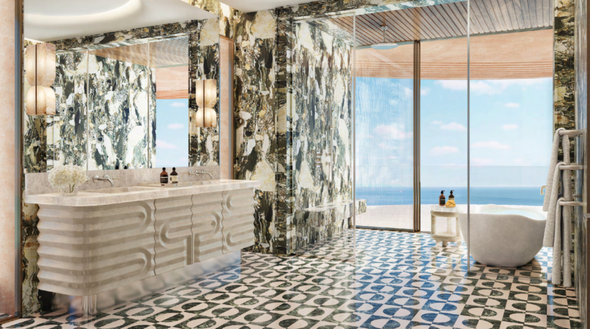 Luxurious bathroom with double washbasin and large wall-mounted mirrors to the left. Marble-patterned walls and geometric tiling on the floor. A freestanding bathtub next to a glass wall overlooks the vast ocean view of Asora Bay under the sunny Dubai sky. Elegant pendant lights hang overhead.