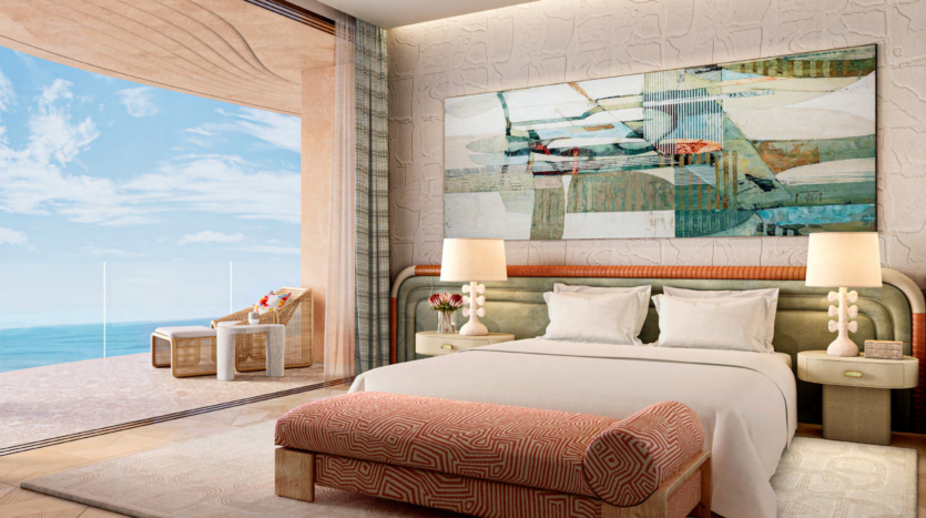 Luxurious room in Asora Bay, Dubai, featuring a king-size bed with decorative pillows and abstract artwork above the headboard. Two unique lamps sit on bedside tables. A glass door opens onto a balcony with wicker chairs and a view of the ocean under a clear blue sky.