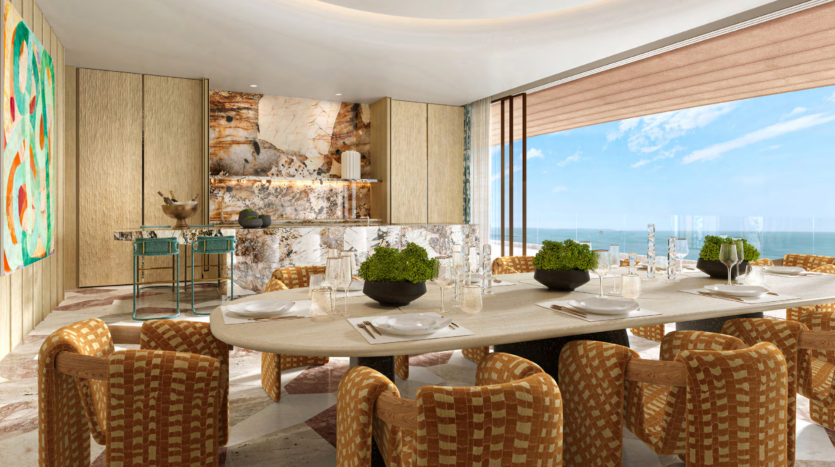 A luxurious dining room with a large table surrounded by wicker chairs overlooks the breathtaking views of Asora Bay. A marble counter and two green stools adorn the background, while a colorful abstract painting adorns the left wall. Large windows frame the breathtaking ocean views, reminiscent of the elegance of Dubai.