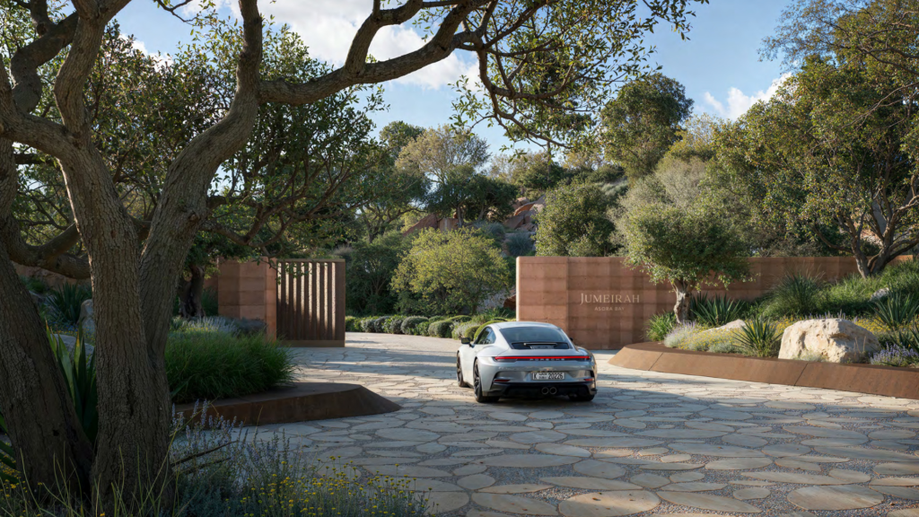 An elegant white car is parked on a patterned stone driveway at Asora Bay, leading to an entrance with large wooden doors. The surrounding area is lush with vegetation and trees, creating a serene atmosphere reminiscent of Dubai's tranquil retreats.