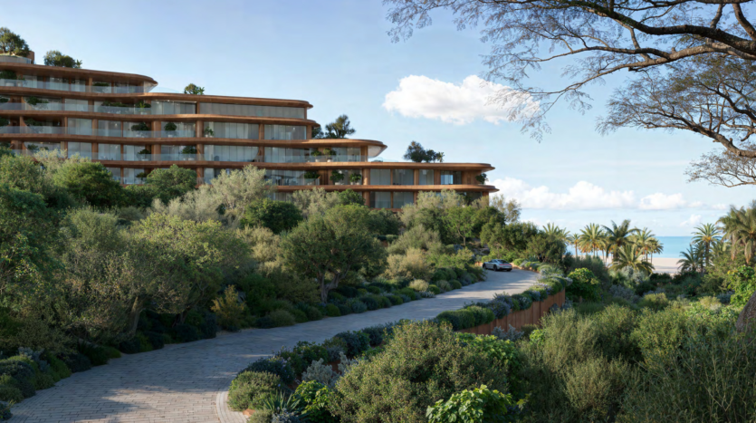 A winding road cuts through lush vegetation and leads to modern multi-storey buildings with large bay windows. Palms and bushes line the path under a clear blue sky, with a glimpse of Dubai's Asora Bay glistening in the background.