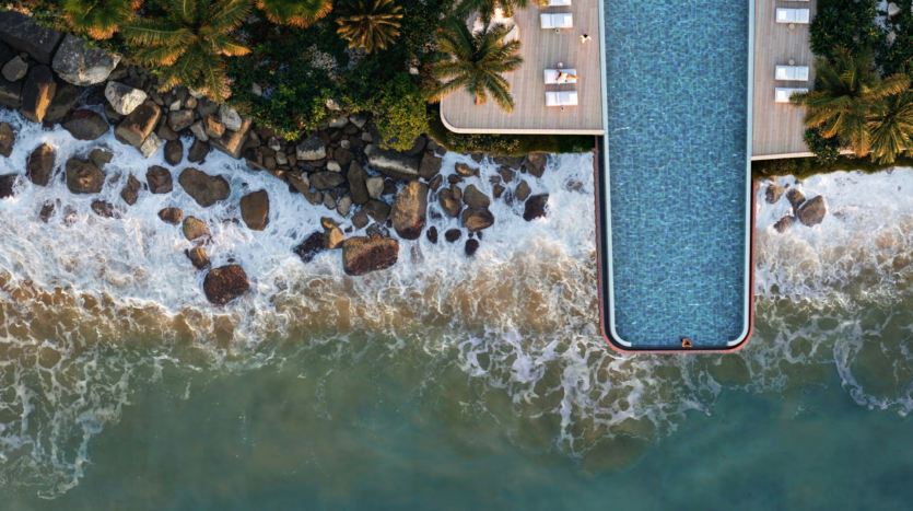 Aerial view of an infinity pool with a swimmer at the edge, facing Dubai's magnificent Asora Bay. The pool is adjacent to a rocky shoreline with waves crashing against the rocks, surrounded by luxurious loungers and swaying palm trees.