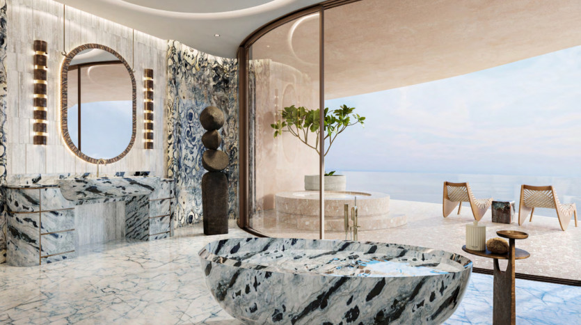 Luxurious bathroom with large curved marble bathtub and matching vanity. A round mirror sits above the vanity. Picture windows reveal a terrace overlooking Dubai's Asora Bay, with two wooden chairs and a small tree, offering ocean views. The marble floor adds elegance.
