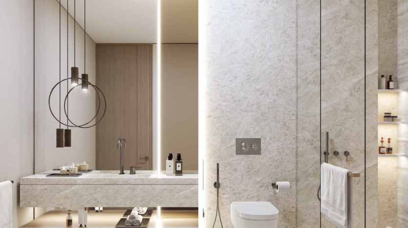 The modern bathroom at Safa Gate Damac features a vertical strip of light that divides the space. To the left, a double washbasin with pendant lights and toiletries completes the minimalist, stone-textured design, while to the right, a toilet and glass-enclosed shower complete this elegant sanctuary.
