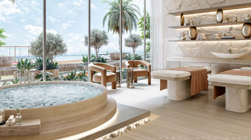 A luxurious spa room with two massage tables, a round Jacuzzi and a lounge area. Large windows reveal sunny exterior views of palm trees and glimpses of the sea from the prestigious Safa Gate Damac. Shelves display decorative objects, reinforcing the serene atmosphere.