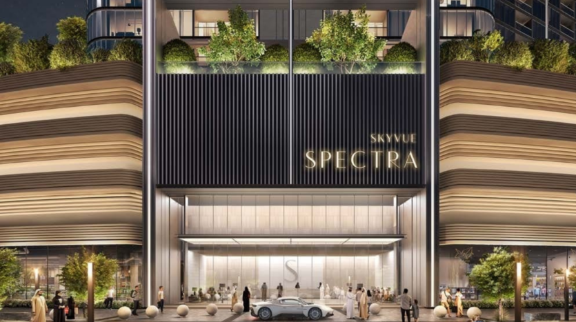 The Skyvue Spectra building, reminiscent of Sobha's architectural elegance, features large glass windows and luminous elements. An elegant car is parked at the entrance, where people gather amidst the greenery-adorned facade, with lights highlighting its superb architectural details.