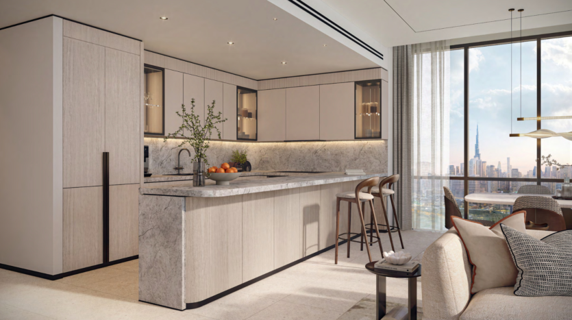 In the heart of the Atelis design district, this modern kitchen features a marble center island with stools, beige cabinets and integrated appliances. A dining table is set nearby, near a large window that reveals a cityscape with a tall tower. Neutral tones and clean lighting complete this contemporary design.